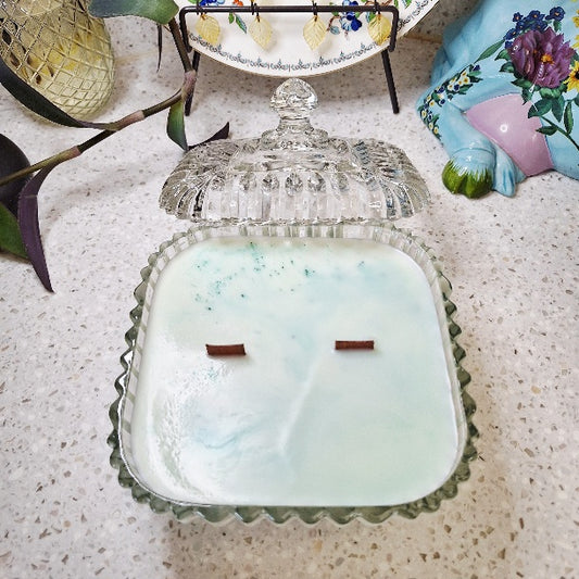 A square glass jar candle with a matching glass lid, filled with pale mint green wax and two wicks, placed on a speckled countertop with decorative objects around it.