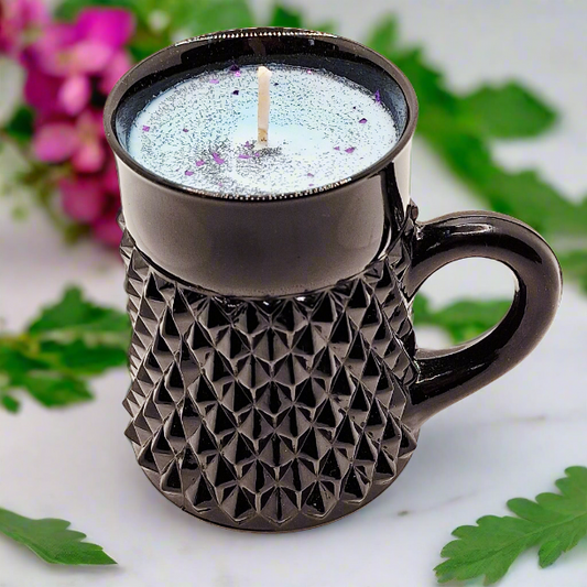 Black textured mug with a candle inside, surrounded by green leaves and pink flowers.