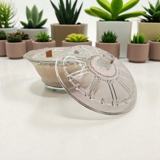 Clear glass jar with a decorative lid on a white surface, with potted plants in the background.