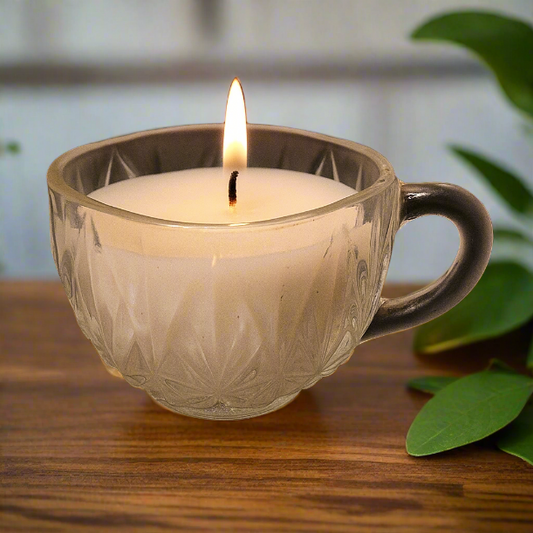 Candle in a glass mug on a wooden surface with green leaves in the background