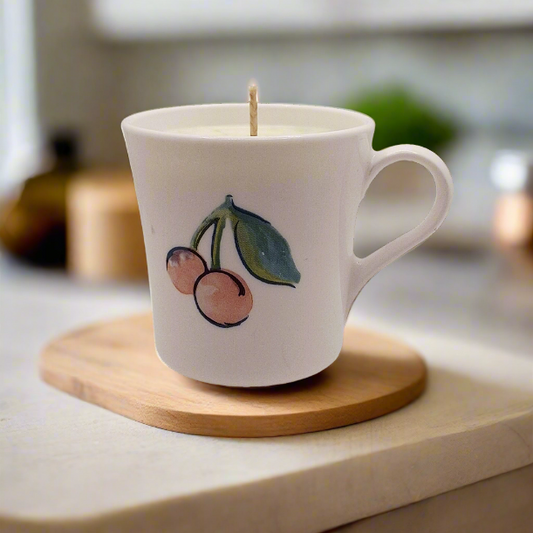 White mug with cherry design containing a candle on a wooden coaster.