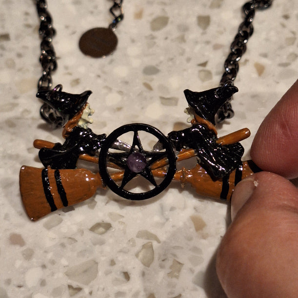 Necklace with witch hat, broomstick, and pentacle pendant held by a hand on a speckled surface.