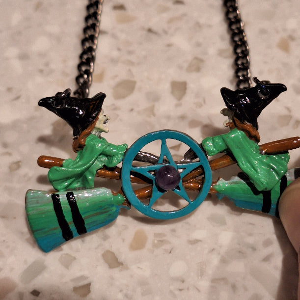 Necklace with witch-themed charms including a broom, hat, and cauldron on a textured surface.