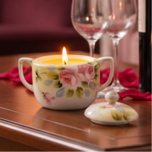Decorative candle in a teacup with floral design on a wooden surface, surrounded by wine glasses and a bottle.
