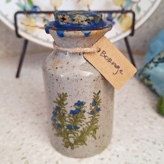 Ceramic bottle with floral design and 'Borage' tag on a textured surface filled with Borage herbs