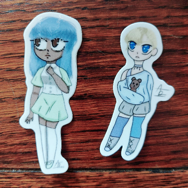 Two die-cut character stickers on a dark wood background. The left sticker features a girl with long blue hair, large glasses, and a light green outfit.