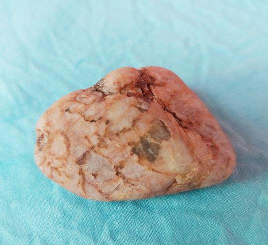 A raw peach moonstone feldspar stone with a pinkish-orange and beige marbled texture, resting on a light blue textured fabric background.