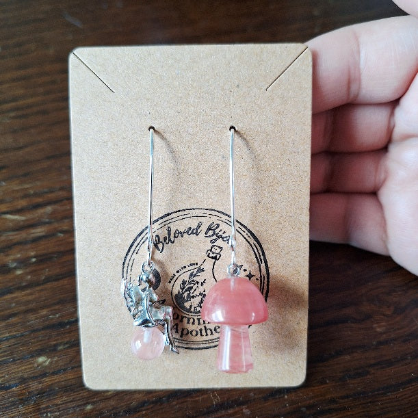Pink mushroom and fairy shaped earrings on a cardboard display card held by a hand.
