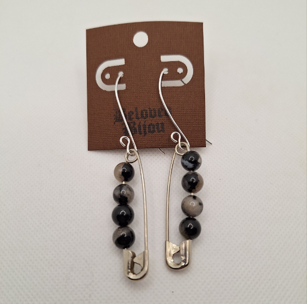 Pair of earrings with safety pins and beaded elements on a branded card against a white background