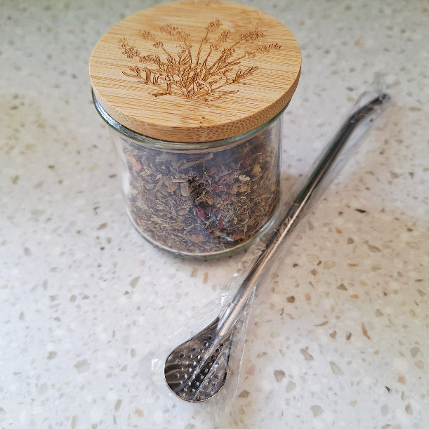 Glass jar with wooden lid containing dried herbs, next to a metal bombilla spoon on a light surface.