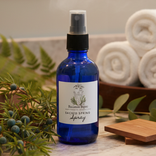 Blue spray bottle labeled 'Sacred Spring Spray' on a marble surface with greenery and towels in the background.