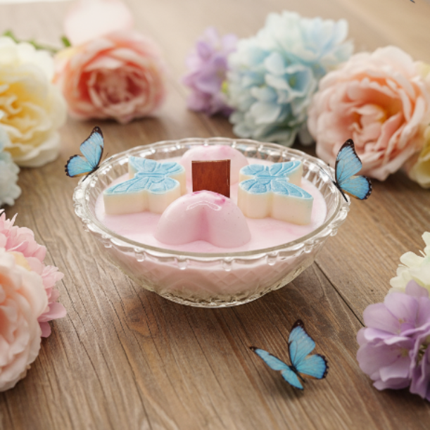 A clear glass bowl filled with pale pink soy wax, topped with three decorative pastel wax butterflies and hearts in white, pink, and light blue, set on a wooden table surrounded by soft pink, peach, and lavender flowers and blue butterflies.