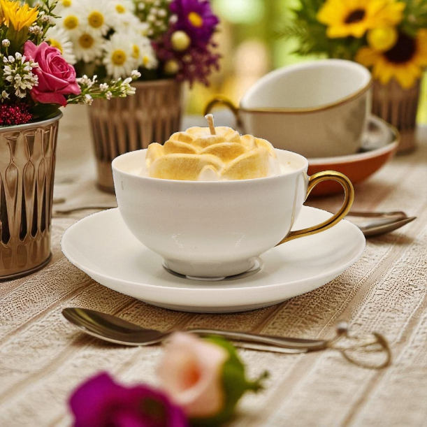 White teacup with a decorative yellow rose candle on a wooden table with flowers and a teapot in the background.
