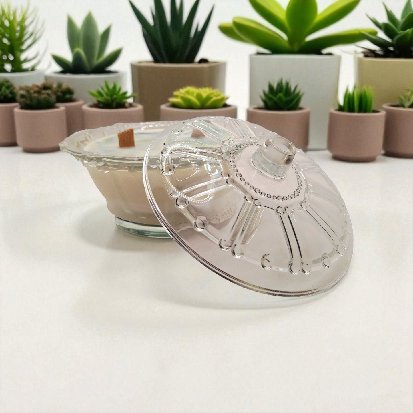 Clear glass jar with a decorative lid on a white surface, with potted plants in the background.