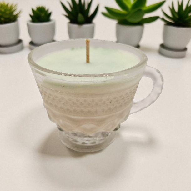 Glass mug candle with a soy wax candle inside on a white surface with potted plants in the background