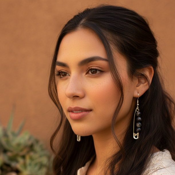 A woman with long dark brown hair wearing a cream-colored embroidered top and silver safety-pin dangle earrings with dark beads, set against a warm, textured stucco wall and a blurred potted plant.