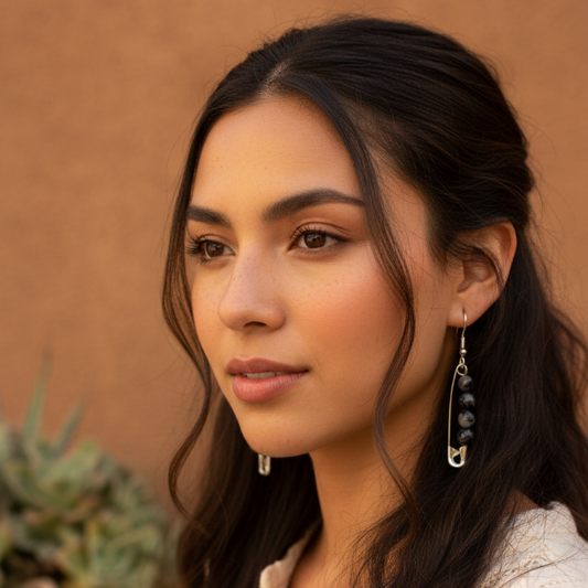 A woman with long dark brown hair wearing a cream-colored embroidered top and silver safety-pin dangle earrings with dark beads, set against a warm, textured stucco wall and a blurred potted plant.